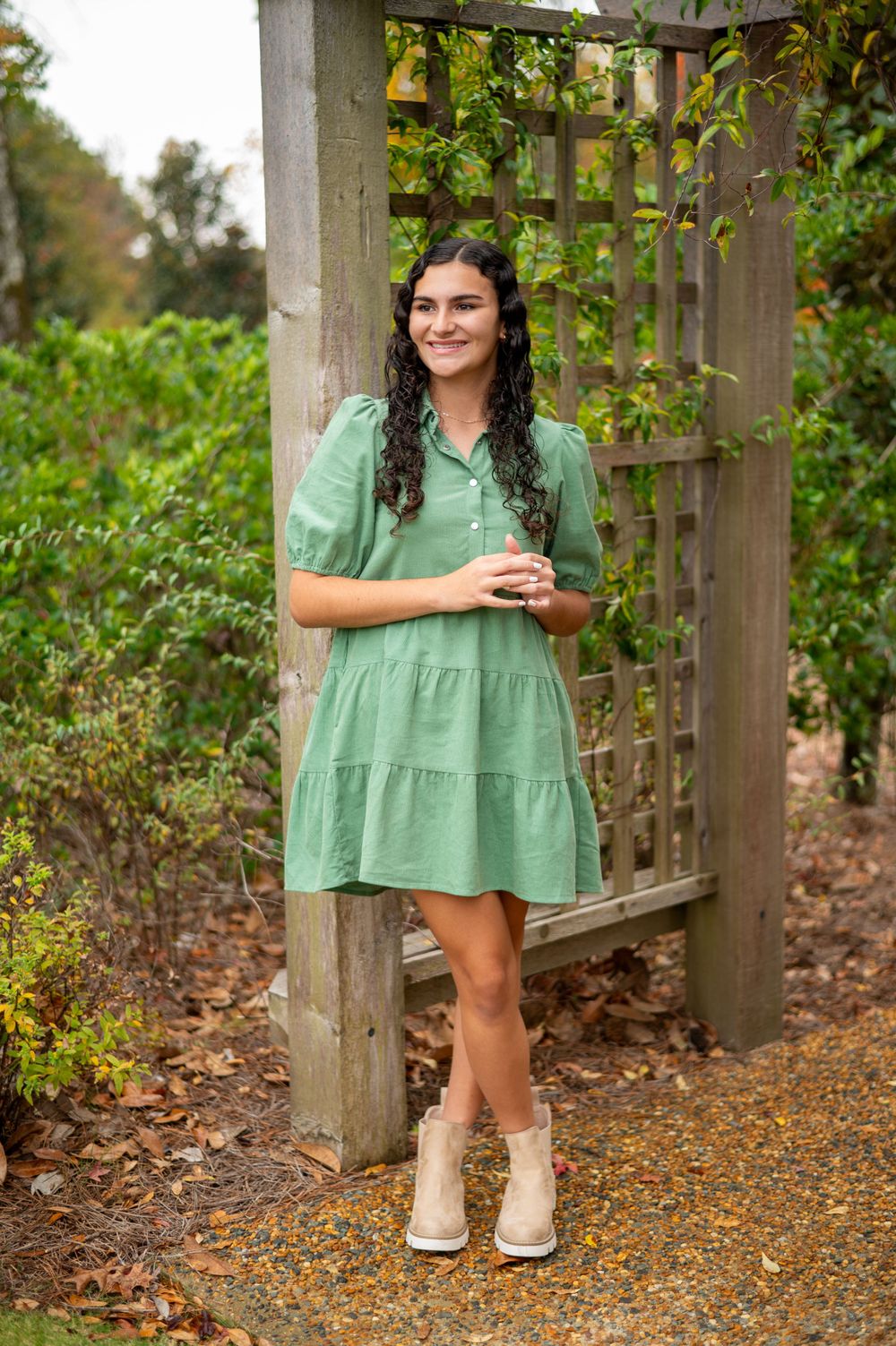 Short sage green tiered dress with boots posed by garden trellis in autumn setting.