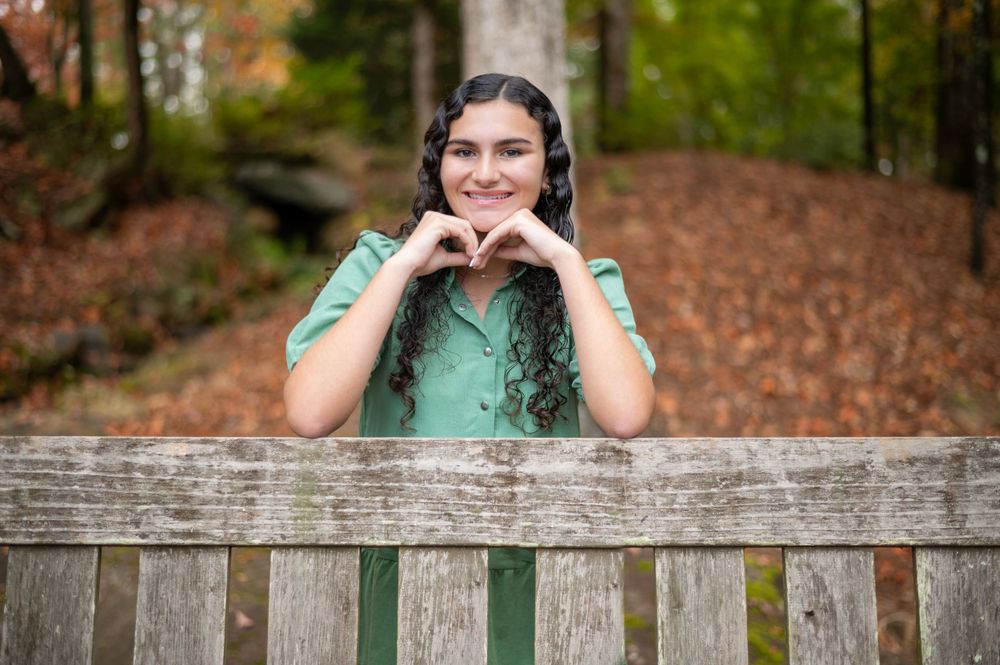 Portrait in sage green dress against wooden bench with autumn foliage backdrop.