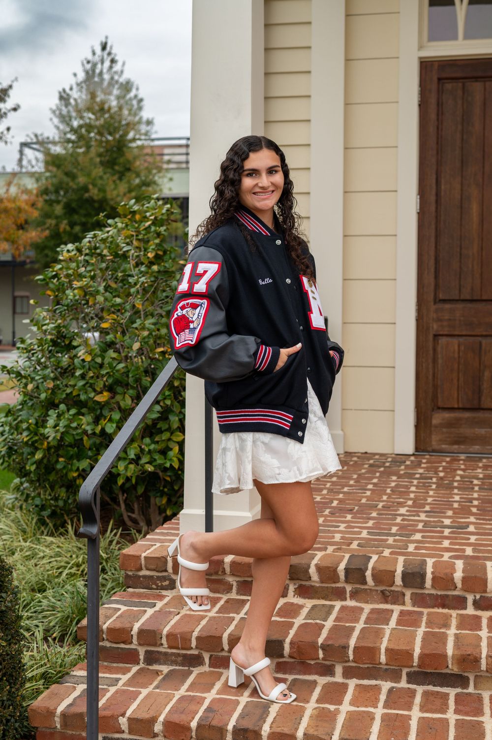 Varsity letterman jacket styled with white dress on brick steps of traditional building.