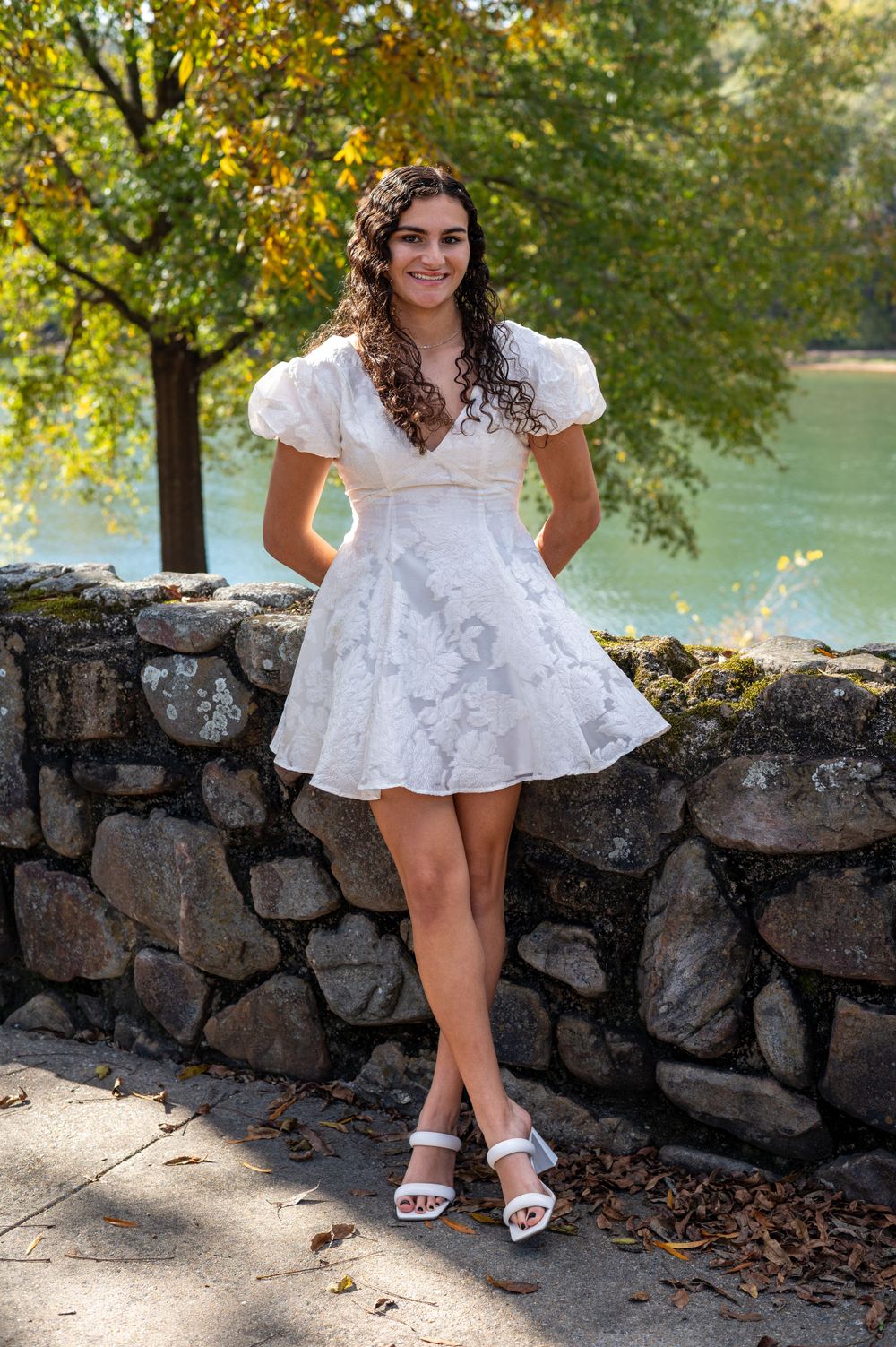Standing by a stone wall in a white dress with autumn foliage in background.