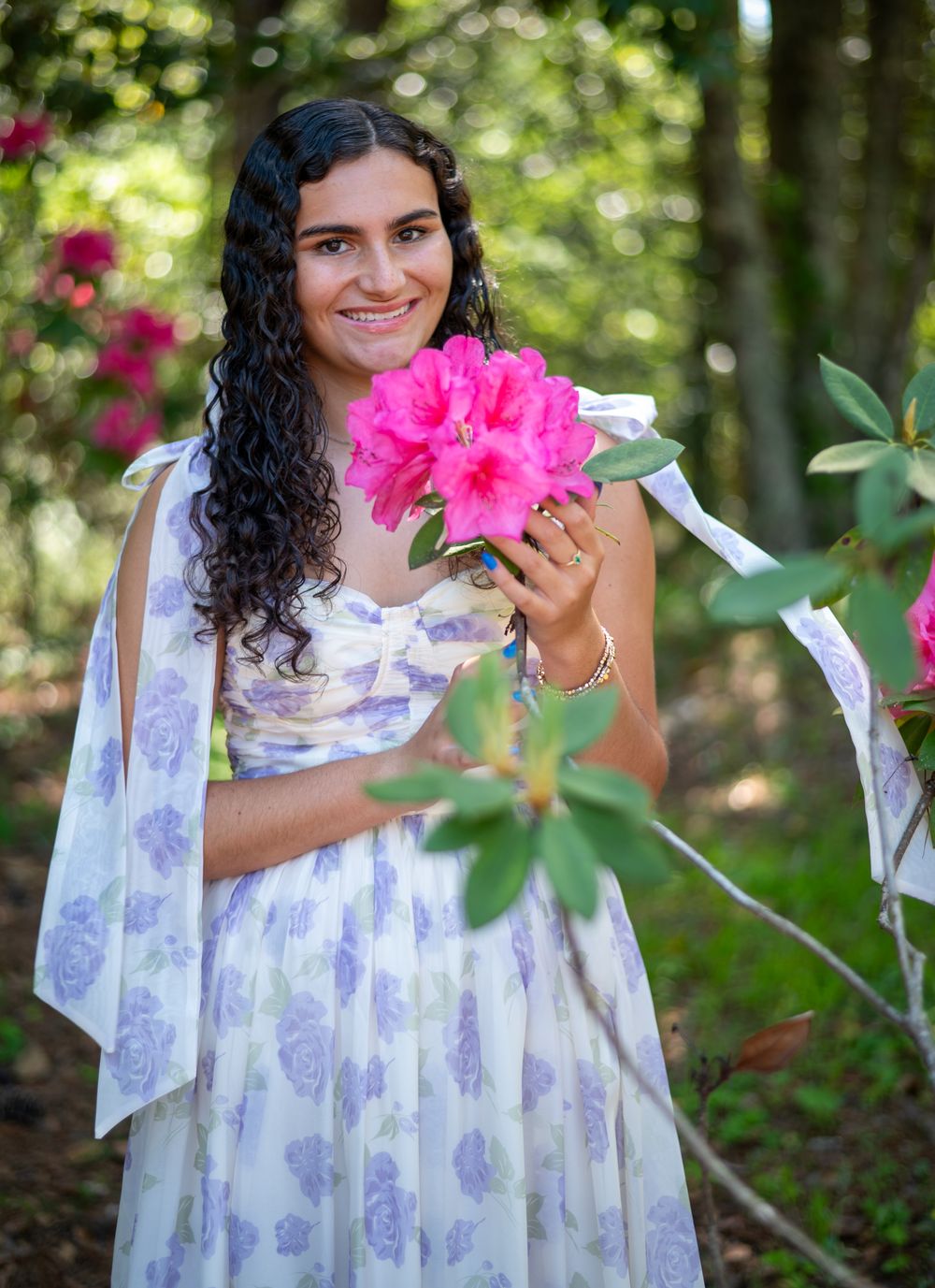 Holding bright pink flowers while wearing a flowing lavender and white floral dress in a garden.