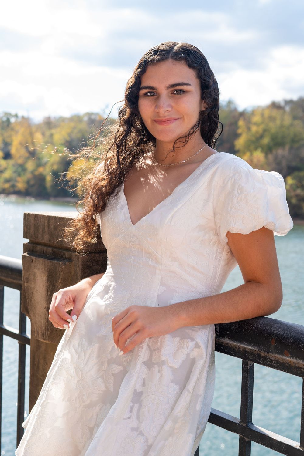 A white summer dress is worn near a waterfront railing with autumn trees in the background.