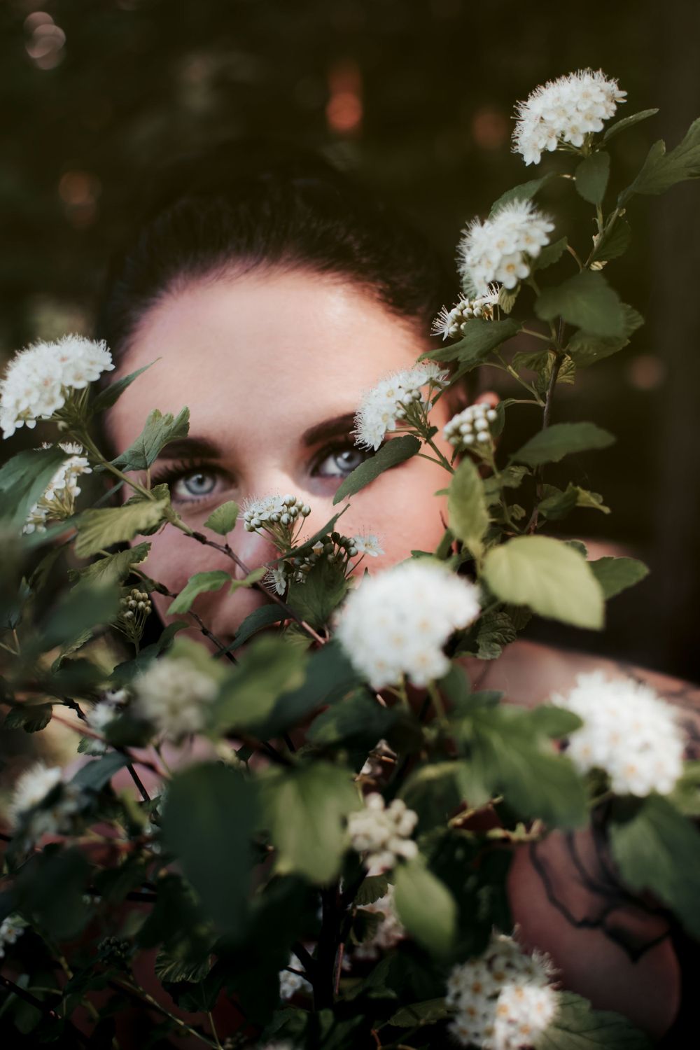 Artistic portrait in Fort Worth, Texas of girl hiding behind puffy white flowers peaking out focusing on her vibrant crystal blue eyes
