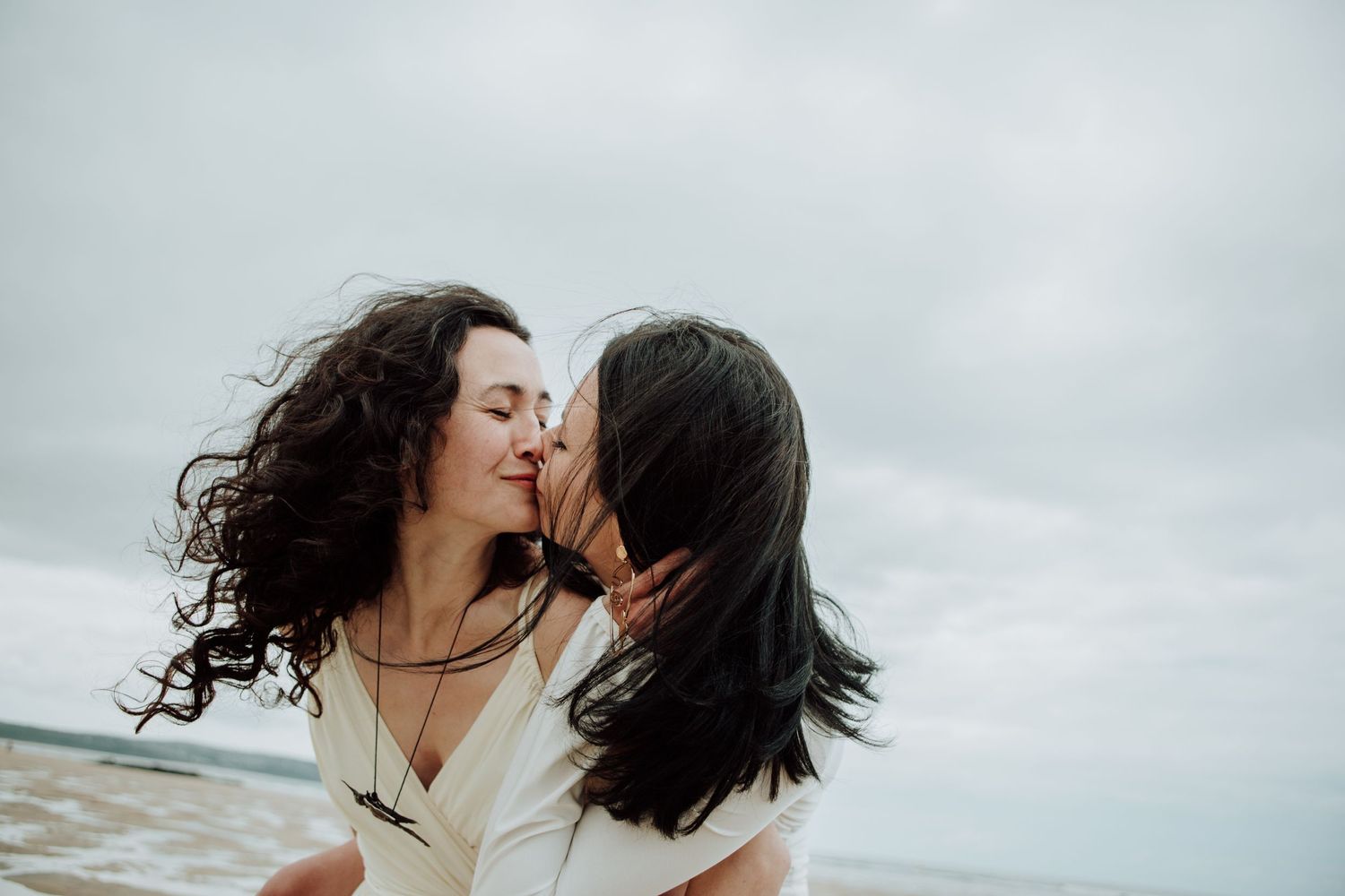 A couple in white dresses share an intimate moment on a cloudy beach.