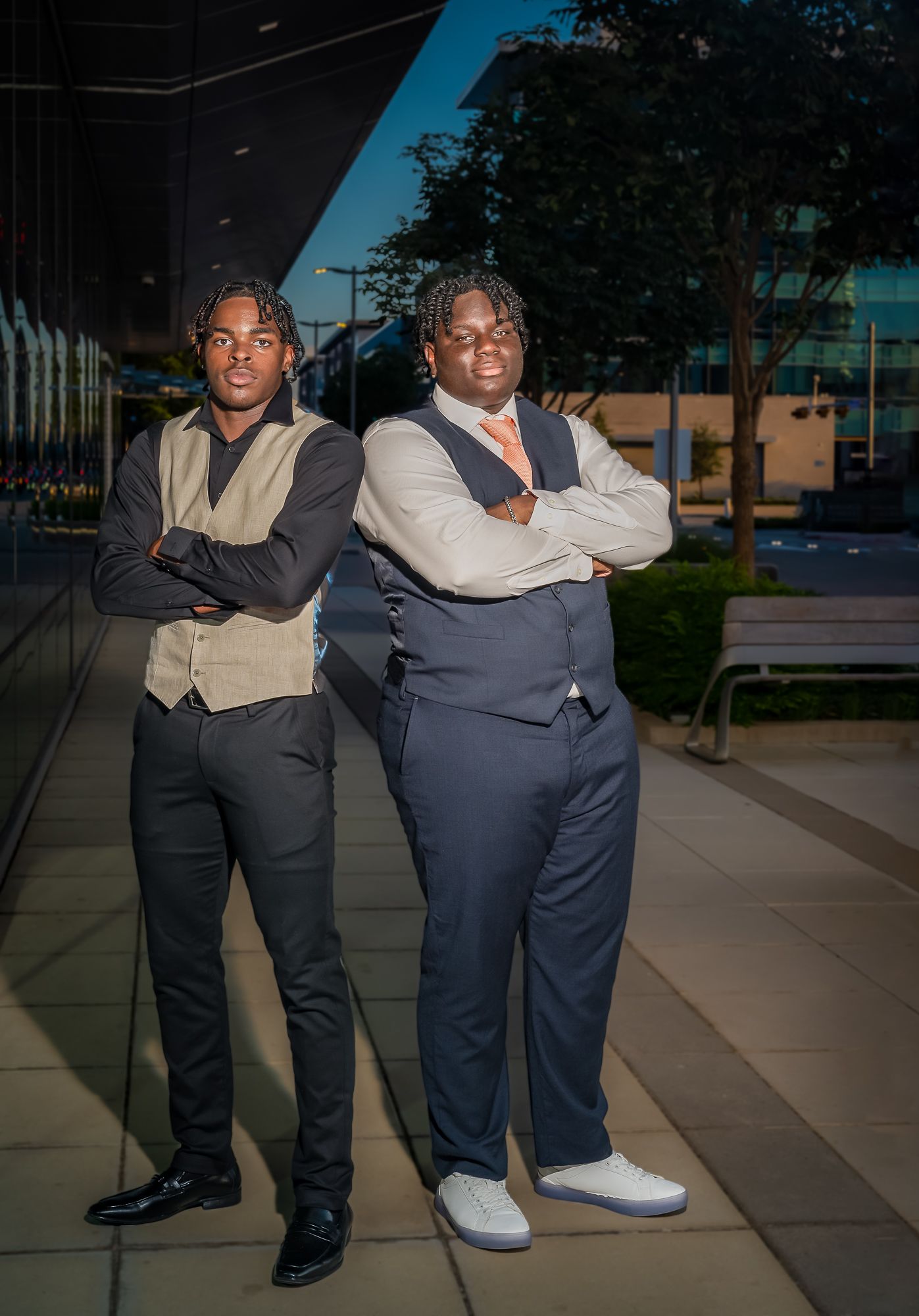 Two friends in formal attire pose confidently against an urban backdrop in evening light.