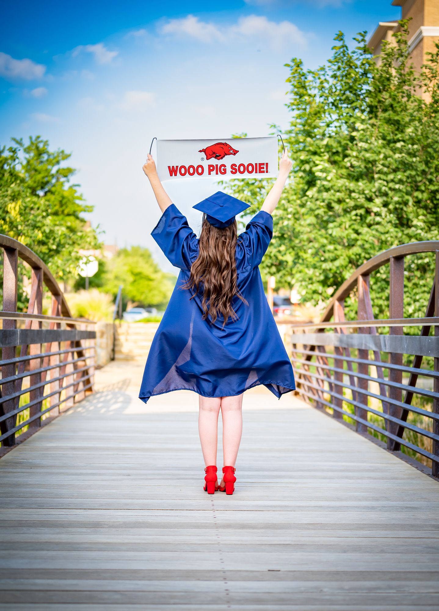 Graduate in blue cap and gown holding Woo Pig Sooie sign on wooden bridge.