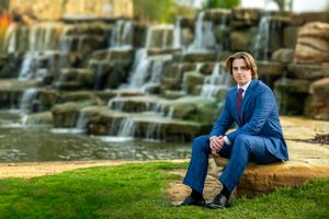 Professional portrait of high school senior in blue suit sitting by cascading waterfall.