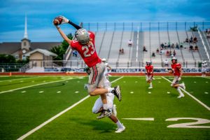 High School Football player in red jersey making impressive leaping catch during game on field. Live action sports photography.