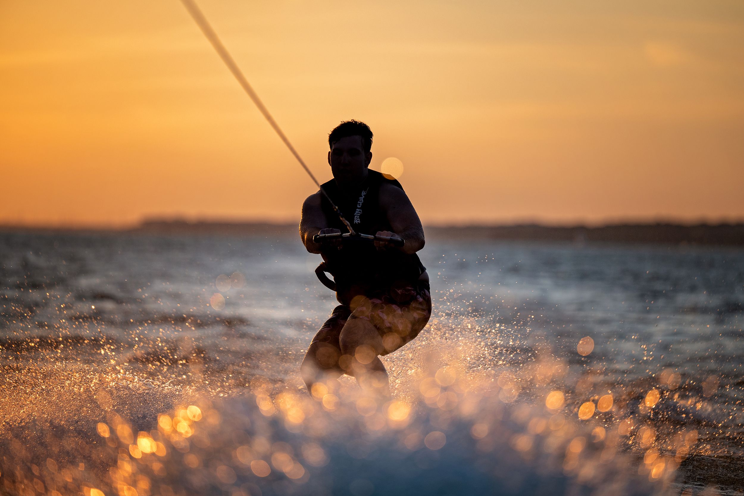 Silhouette of a wakeboarder riding ocean waves at golden sunset, water splashing dramatically in warm orange light.