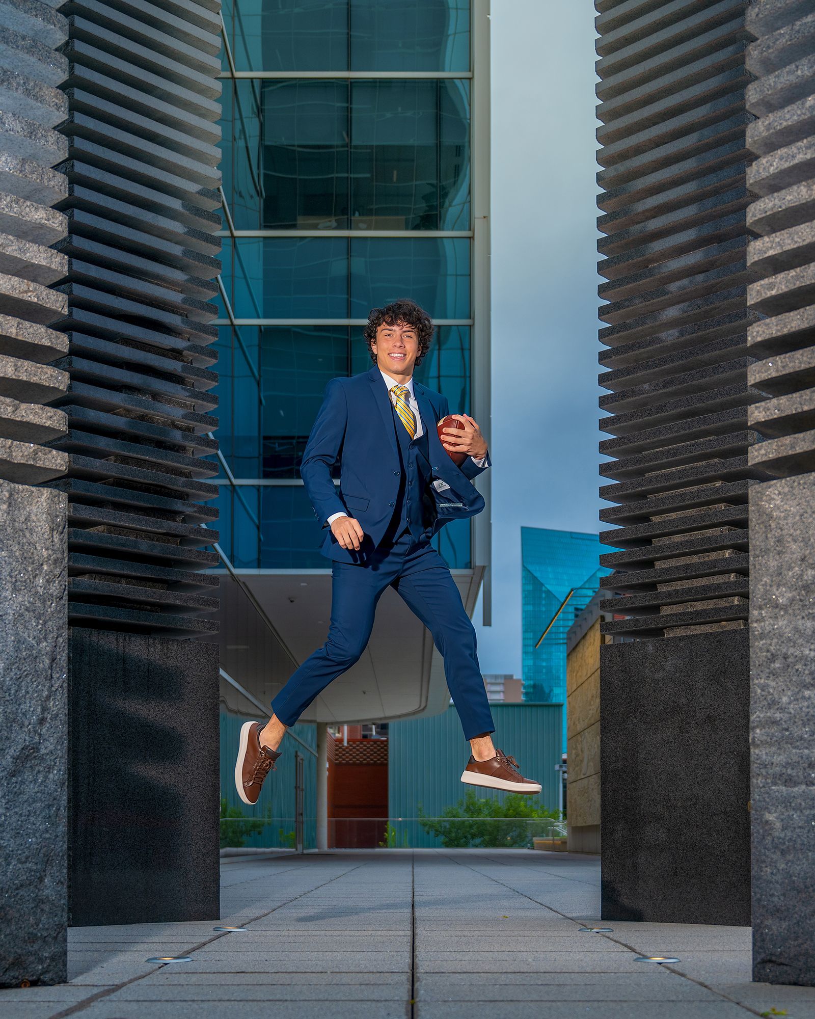 Young man in navy blue suit jumping mid-air between modern architectural columns outdoors.