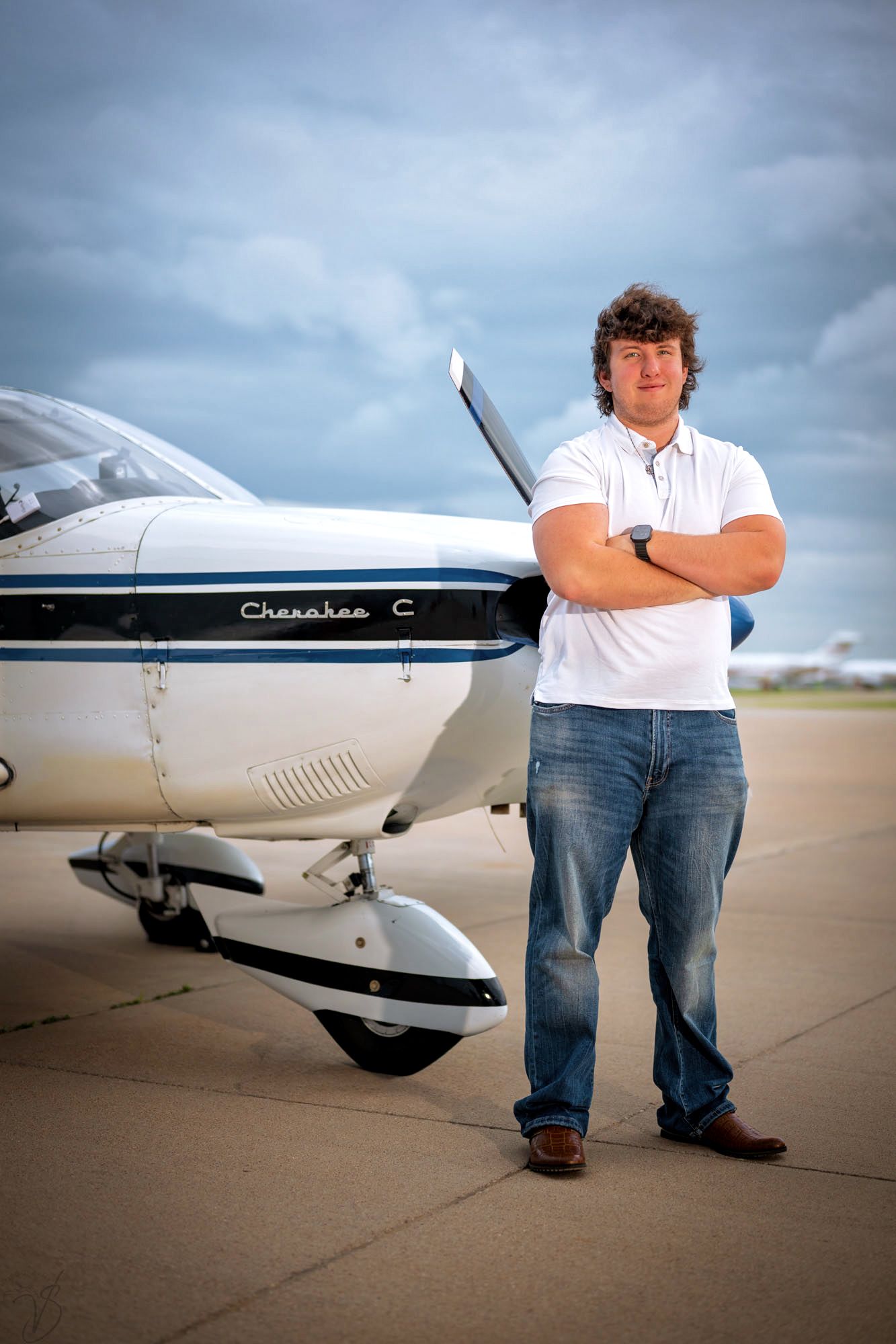 Young man in white polo and jeans stands arms crossed beside a Piper Cherokee C small aircraft on tarmac.