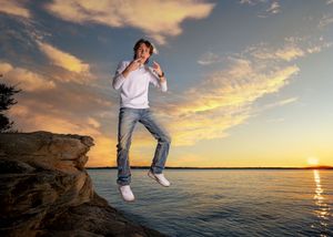 Young man jumping on rocky shoreline at sunset with colorful sky and calm water in background.