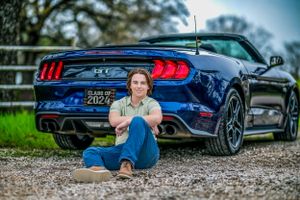 Dark blue Ford Mustang sports car parked on gravel with high school senior seated casually next to rear bumper.