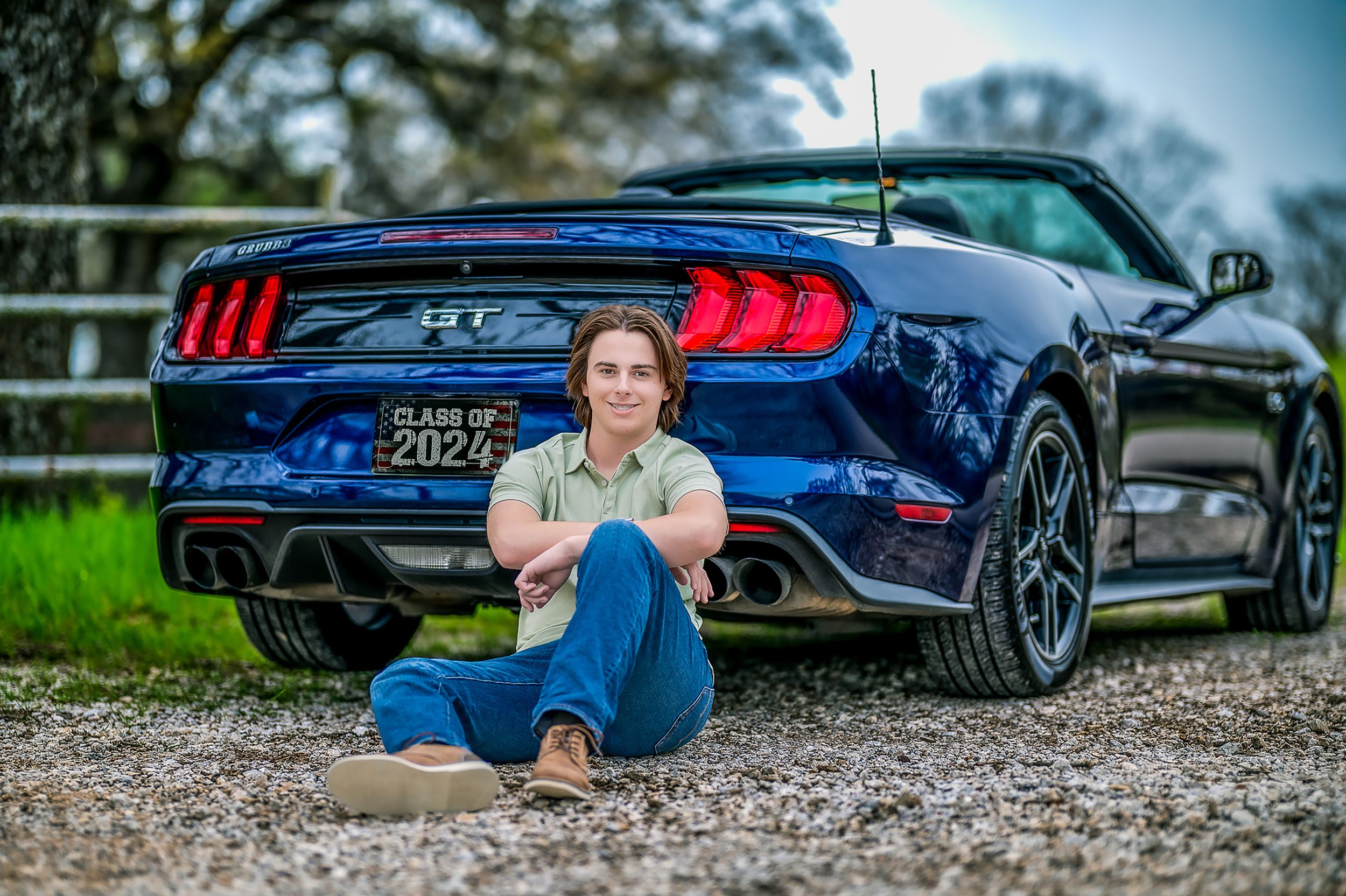 Dark blue Ford Mustang sports car parked on gravel with high school senior seated casually next to rear bumper.