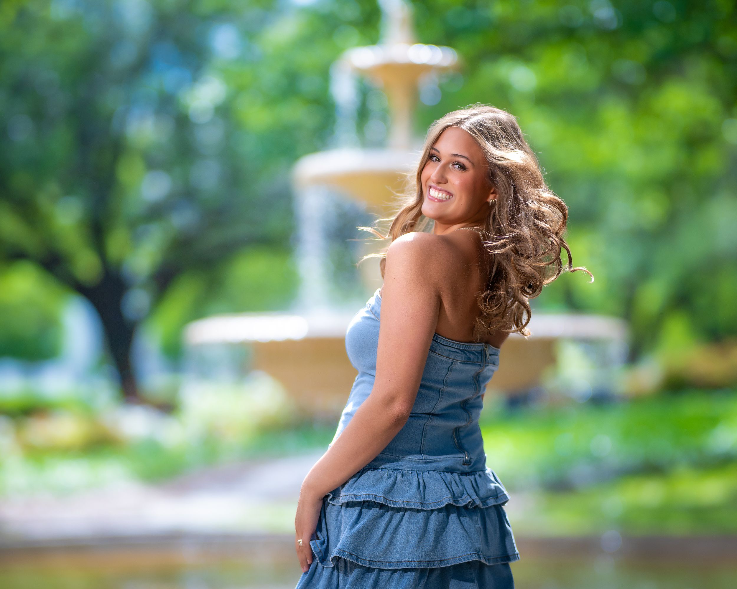 Young woman in blue ruffled dress smiling over shoulder near garden fountain with lush green trees.