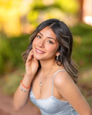 Young woman smiling outdoors, wearing a silver top and layered necklaces, with soft bokeh green background.