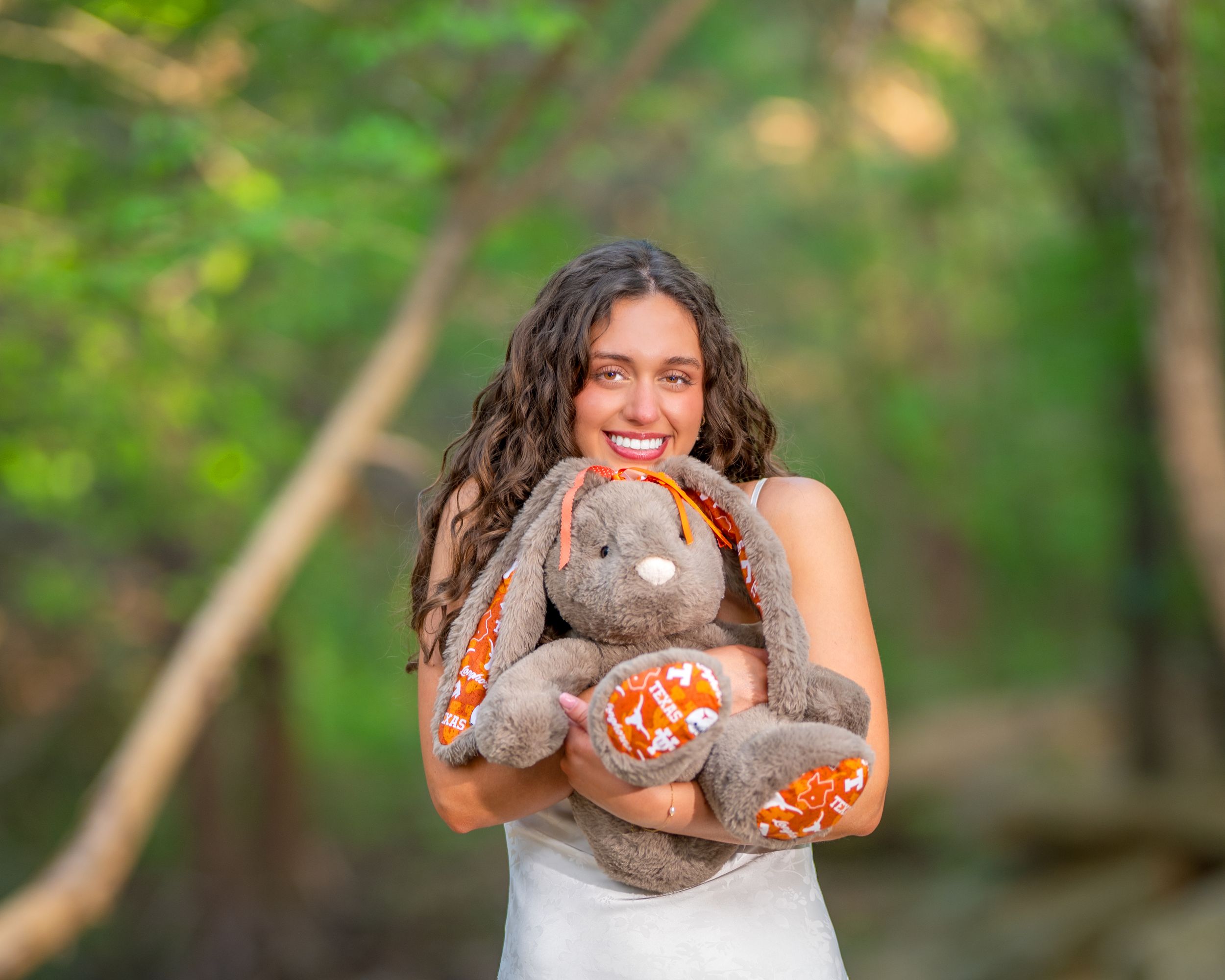Smiling young woman in white dress hugging a large brown and orange stuffed bunny rabbit outdoors in a green forest.