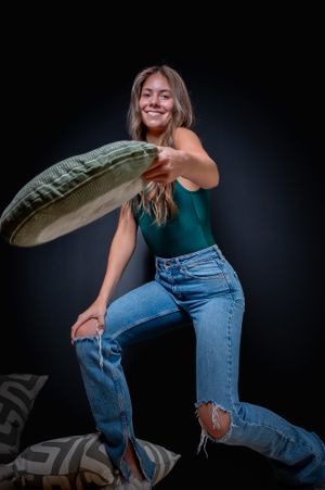 A person in ripped jeans and green top poses playfully with a large cushion against a dark background.