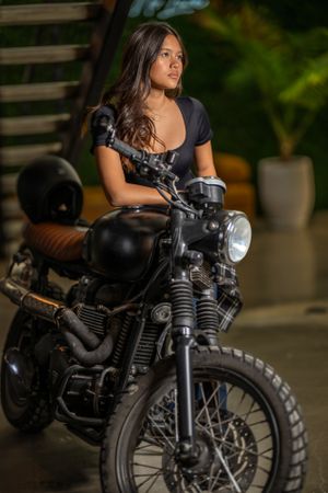 A teen sitting on a vintage motorcycle in moody lighting with a classic headlight.
