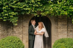 Bride and groom share a romantic moment under ivy-covered stone archway with manicured hedges at elegant wedding venue.