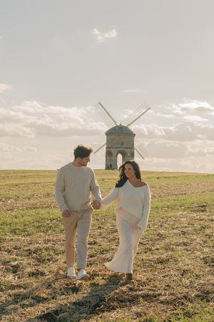 A couple in white and gray outfits walks together across a sunlit grassy meadow on a cloudy day.