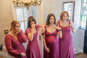 Four bridesmaids in mauve gowns share an emotional moment, one visibly pregnant, reacting with joy and tears.