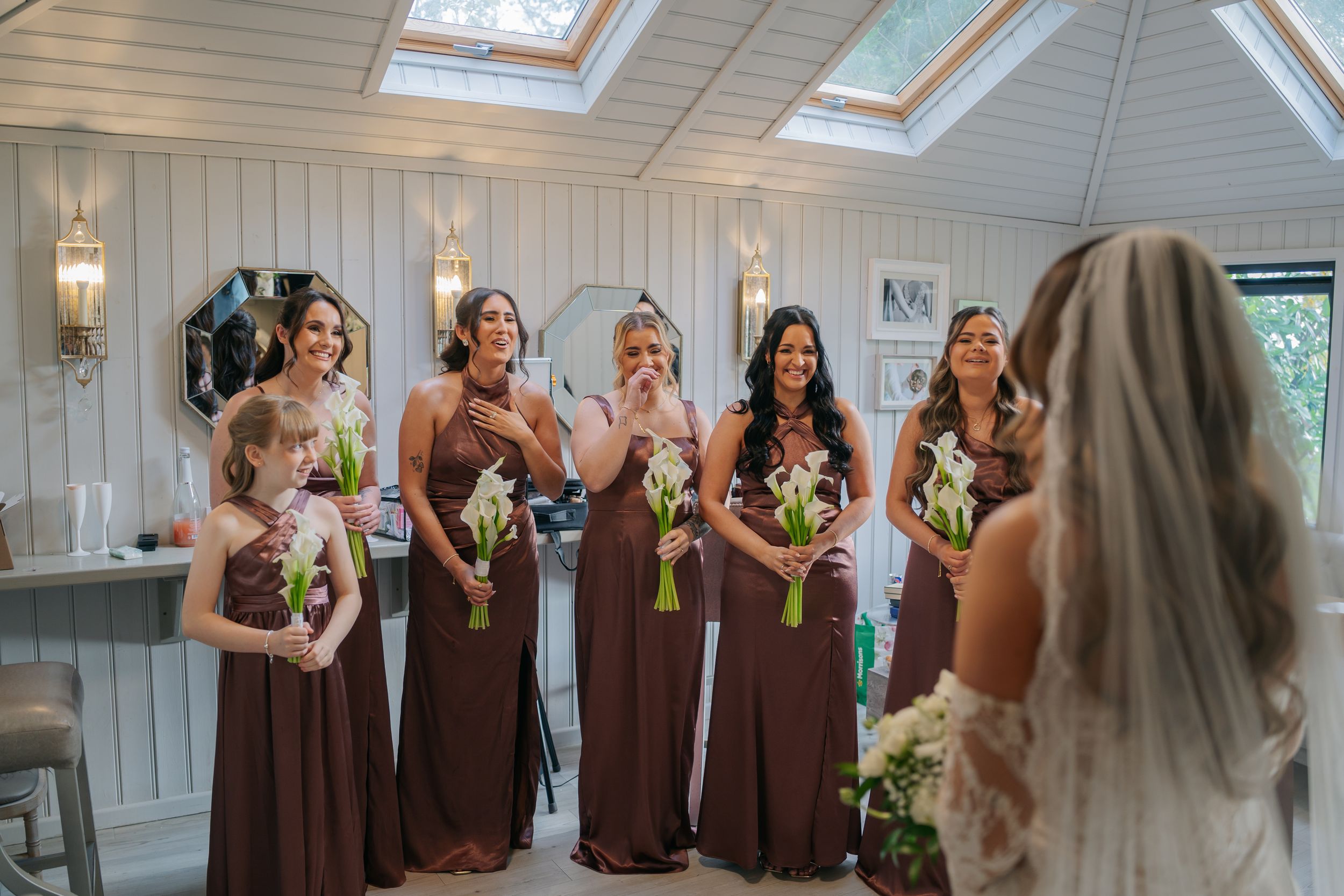 A wedding party in brown dresses reacts with excitement and joy in a bright white room with skylights.