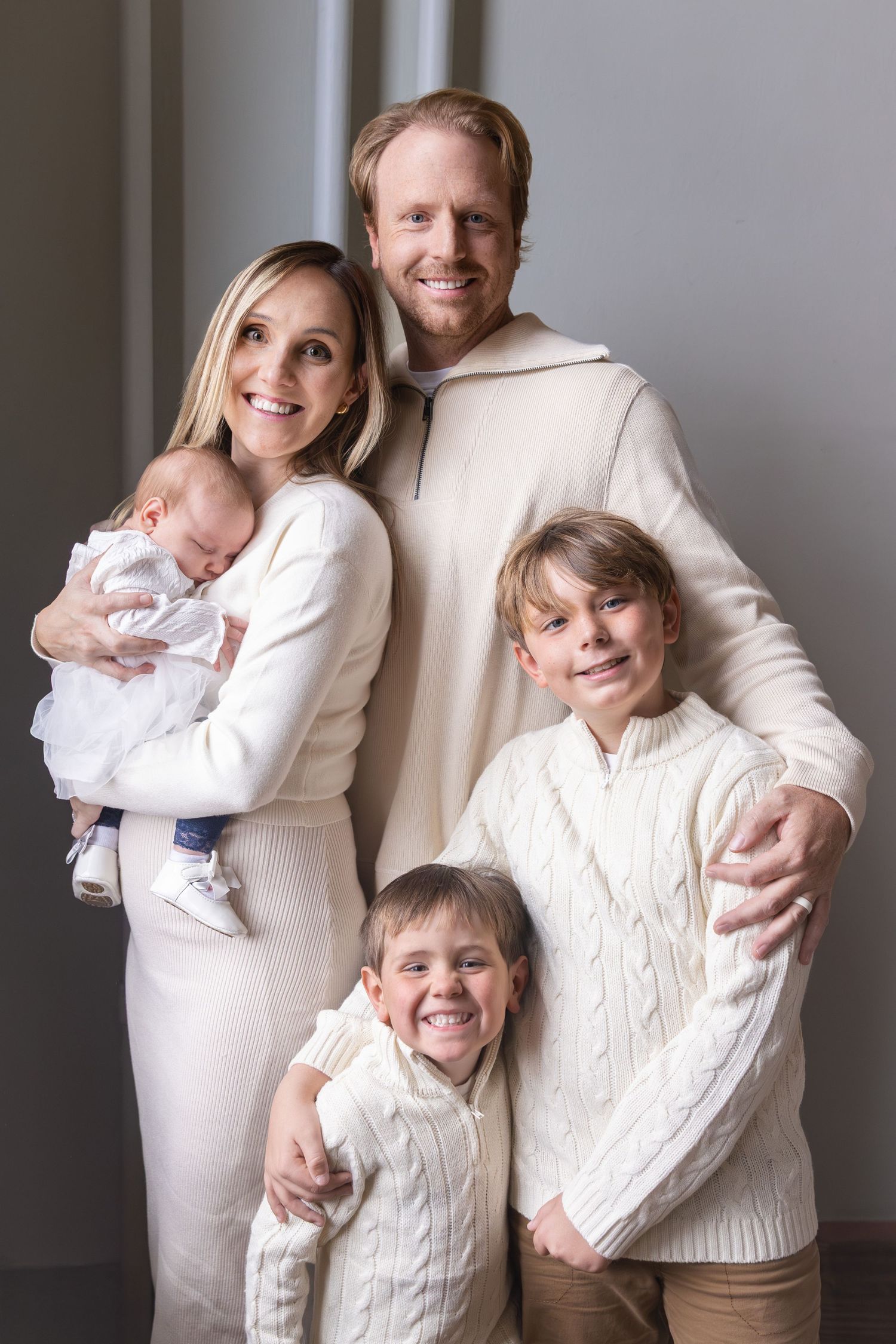 Family portrait in coordinated white and cream outfits against a neutral wall.