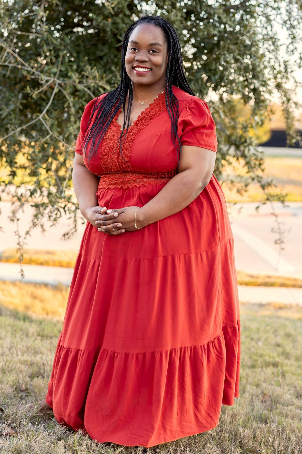 Woman in flowing red maxi dress with lace trim poses outdoors at sunset in grassy field.