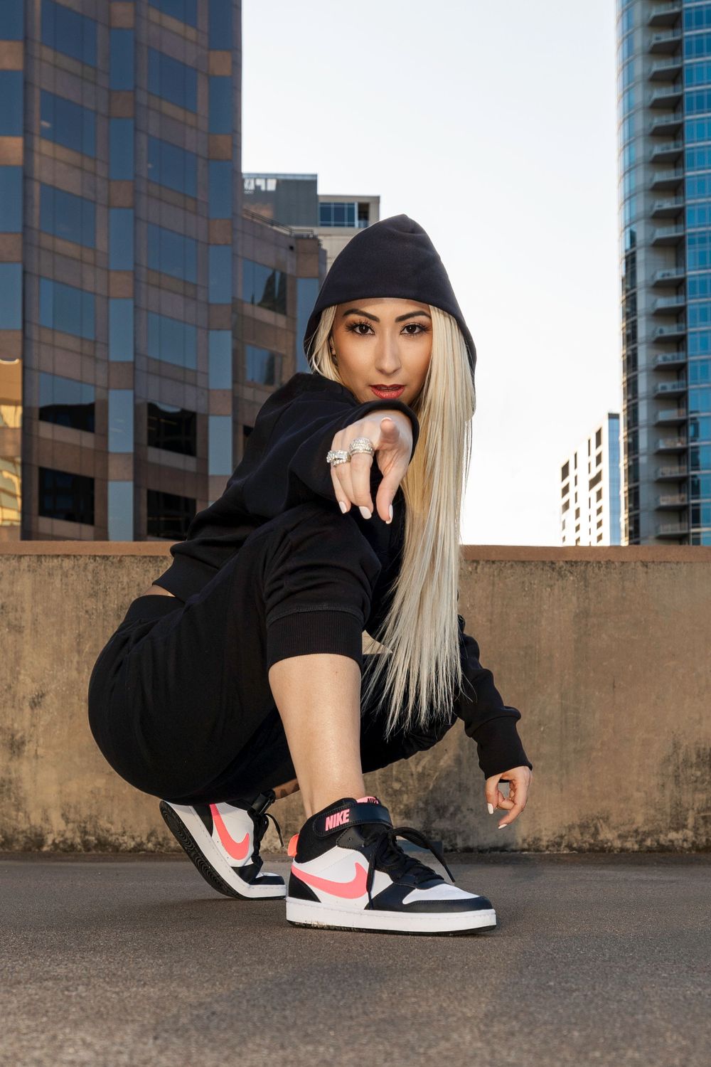 A person in a black hoodie and blonde hair poses on the ground with Nike sneakers against an urban backdrop.