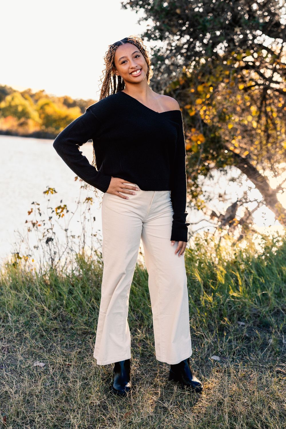 A person wearing a black top and white pants poses outdoors during golden hour near tall grass and water.