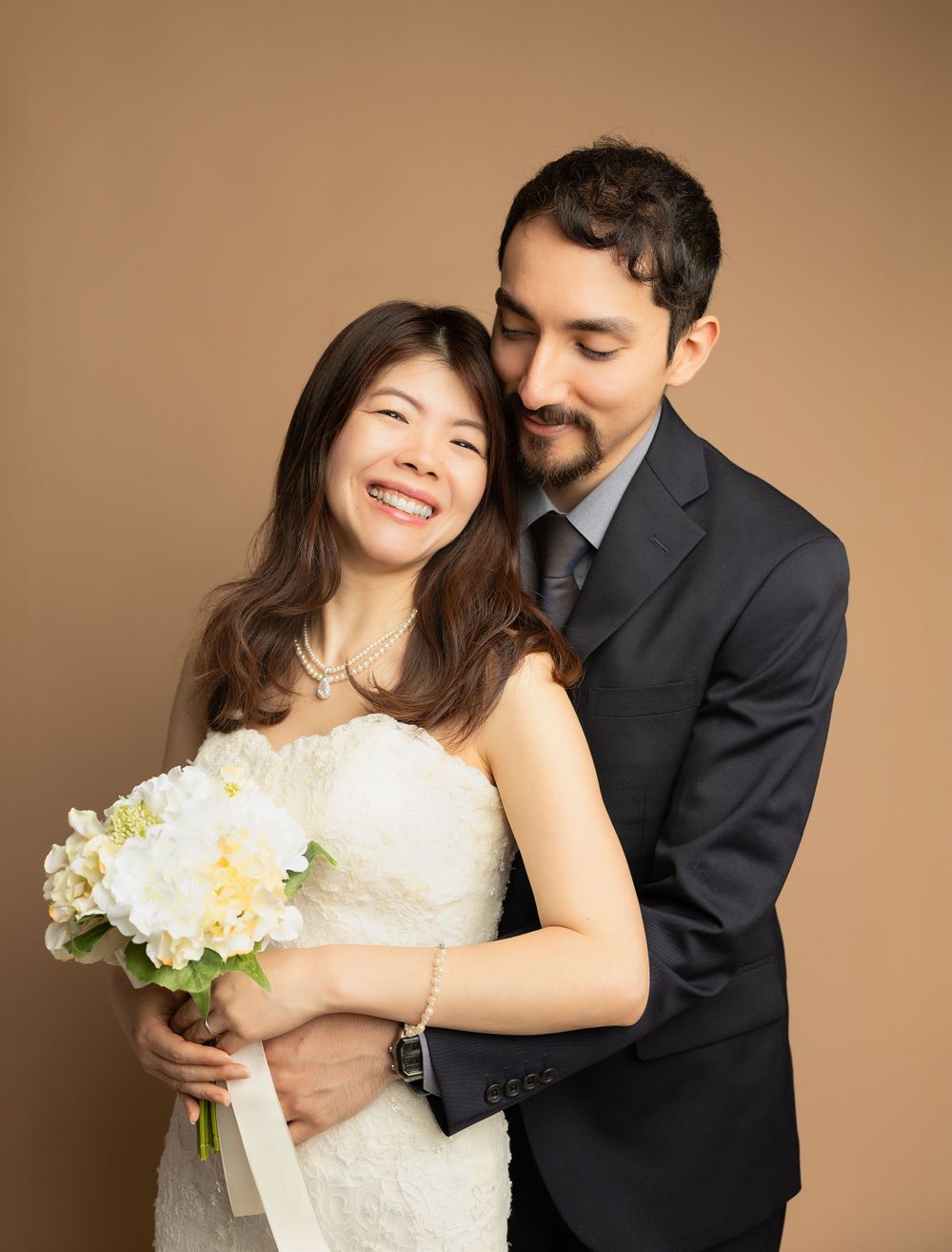 Studio portrait of couple in wedding attire with white bouquet against brown background.