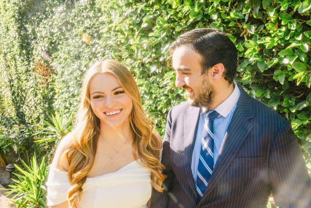 Couple smiles radiantly in formal attire against lush green garden backdrop.