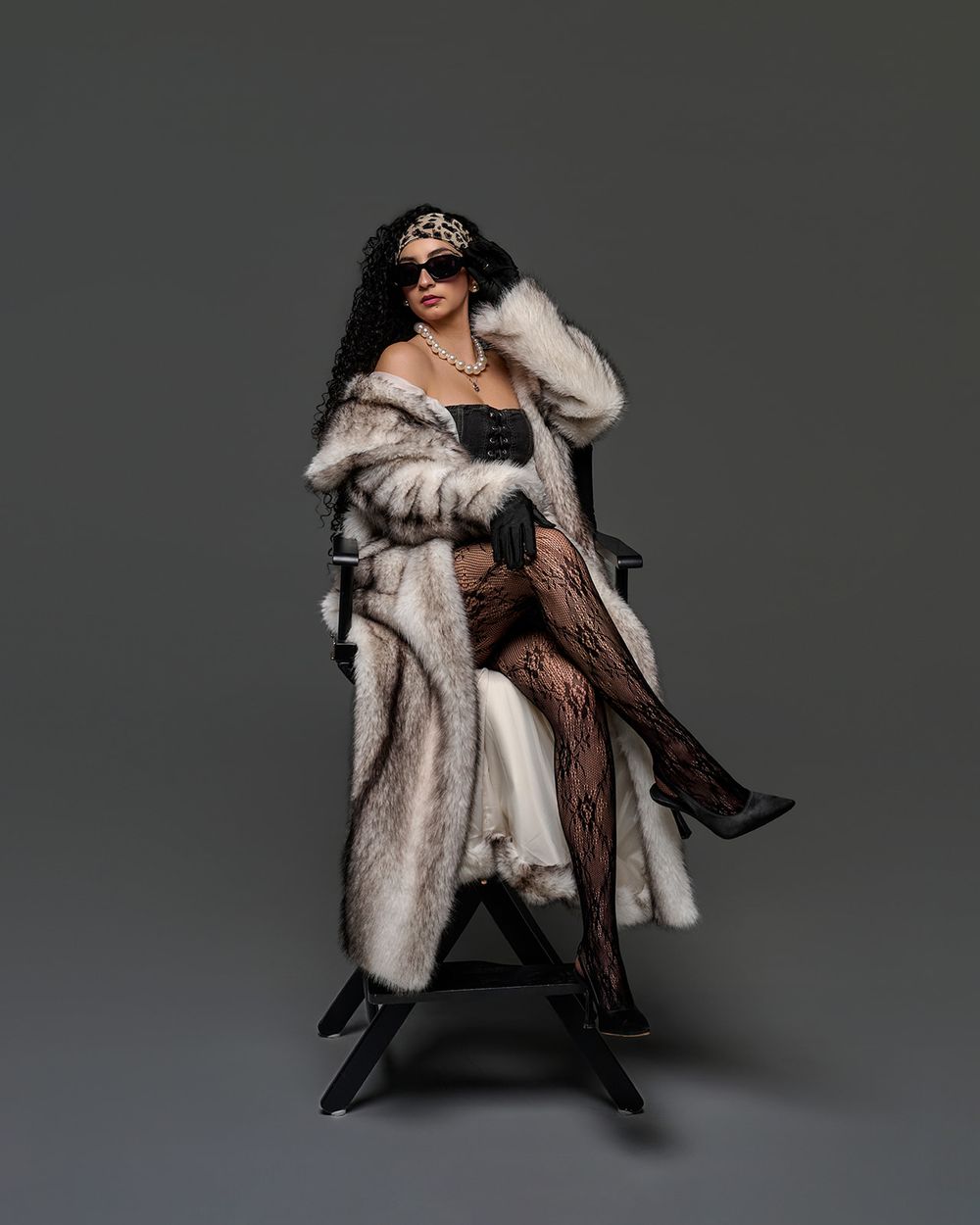 A model poses dramatically in a black chair wearing a luxurious white fur coat and dark evening attire against a gray backdrop.