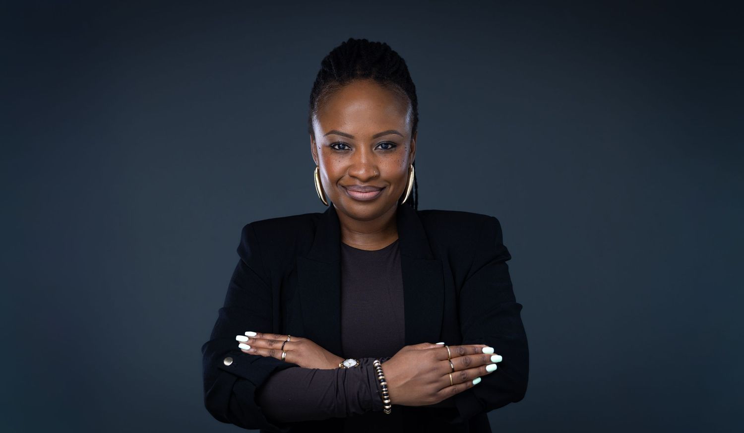 A person in a black blazer wearing a dramatic white shell statement necklace poses with raised hands against dark background.