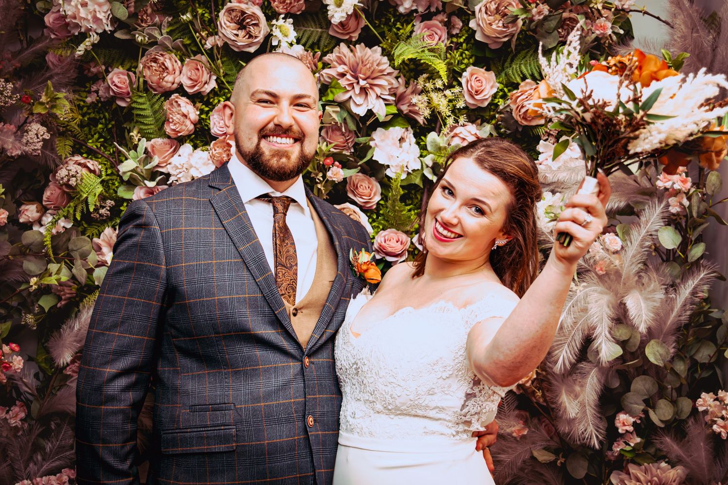 Newlyweds celebrating joyfully in front of pink floral wall backdrop.