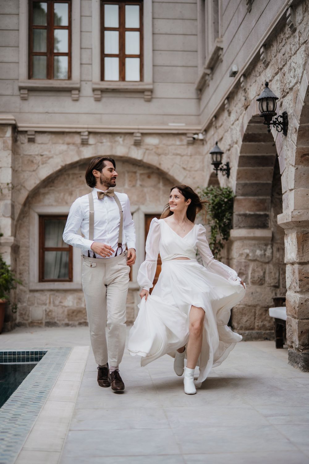 A couple in wedding attire playfully walks together through a historic stone courtyard with elegant architecture.