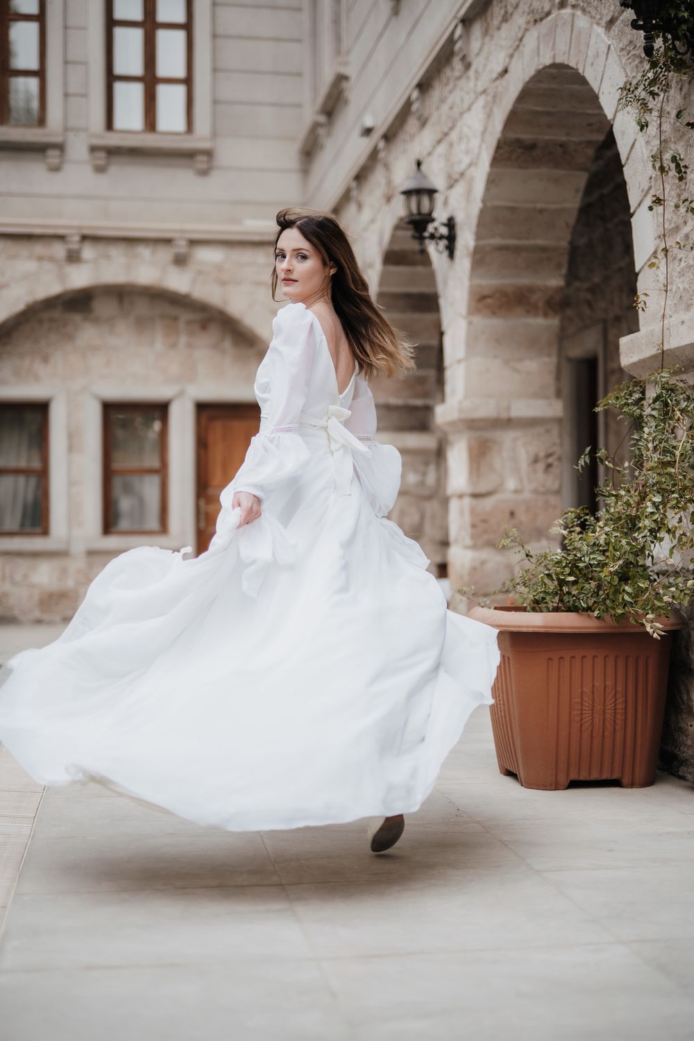 A graceful woman in a flowing white dress twirls beneath stone arches of a classical building.