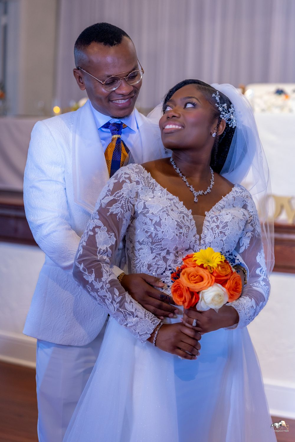 A bride holding orange and red bouquet poses with groom in white suit and blue tie.