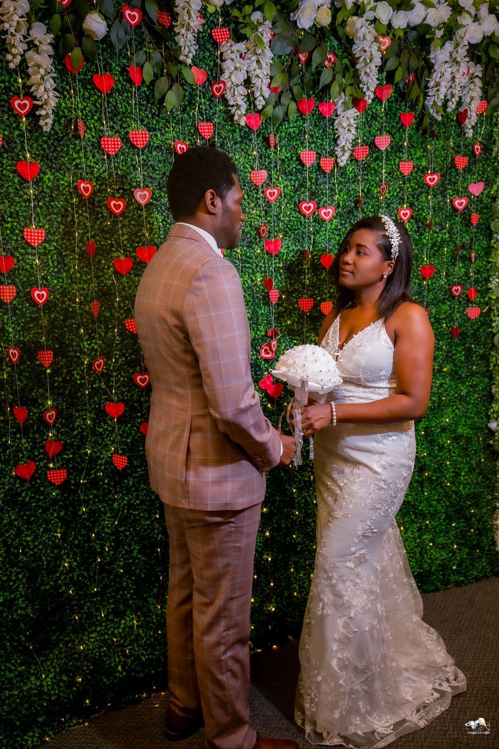A couple shares an intimate moment during their wedding against a wall decorated with red hearts.