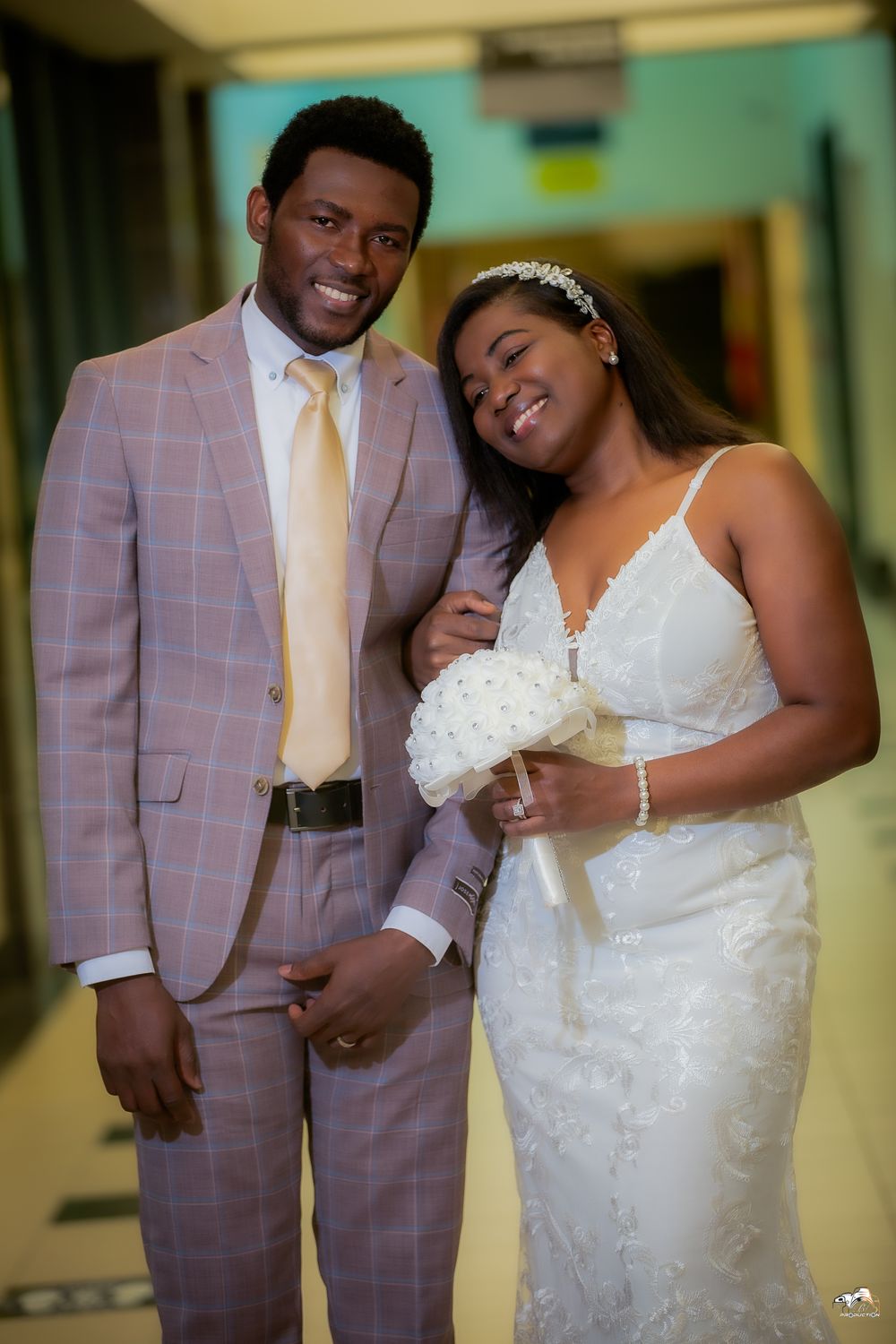 A happy newlywed couple poses in a hallway, groom in pink suit and bride in white dress.