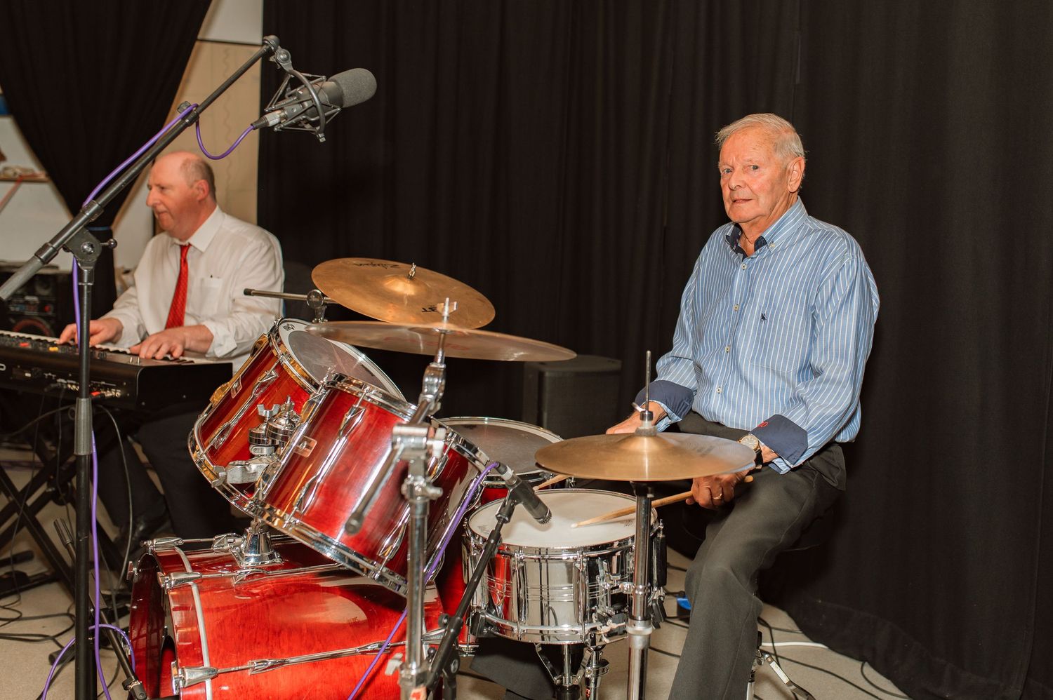 Two musicians performing on bright red drum sets against a black curtain background during a concert.