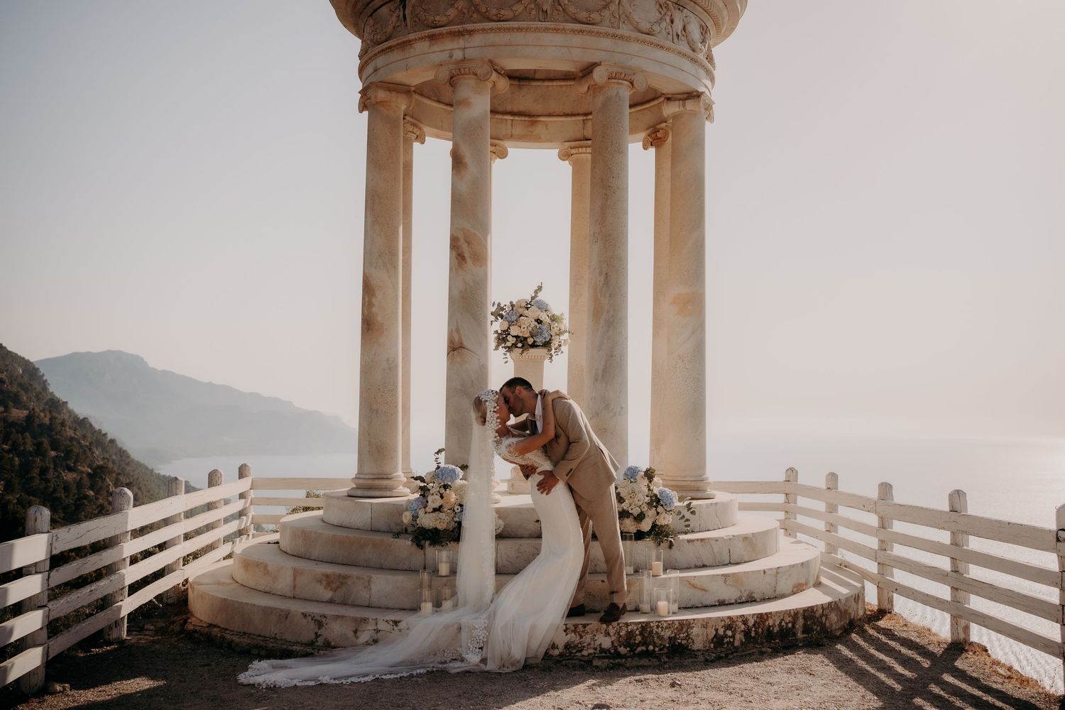 A romantic moment captured at a classical columned gazebo overlooking a misty mountain landscape with white railings.