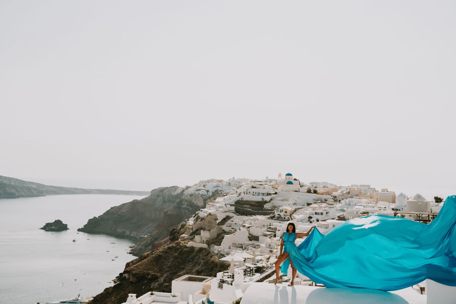 Flowing turquoise fabric billowing against Santorini's coastal landscape and white buildings.