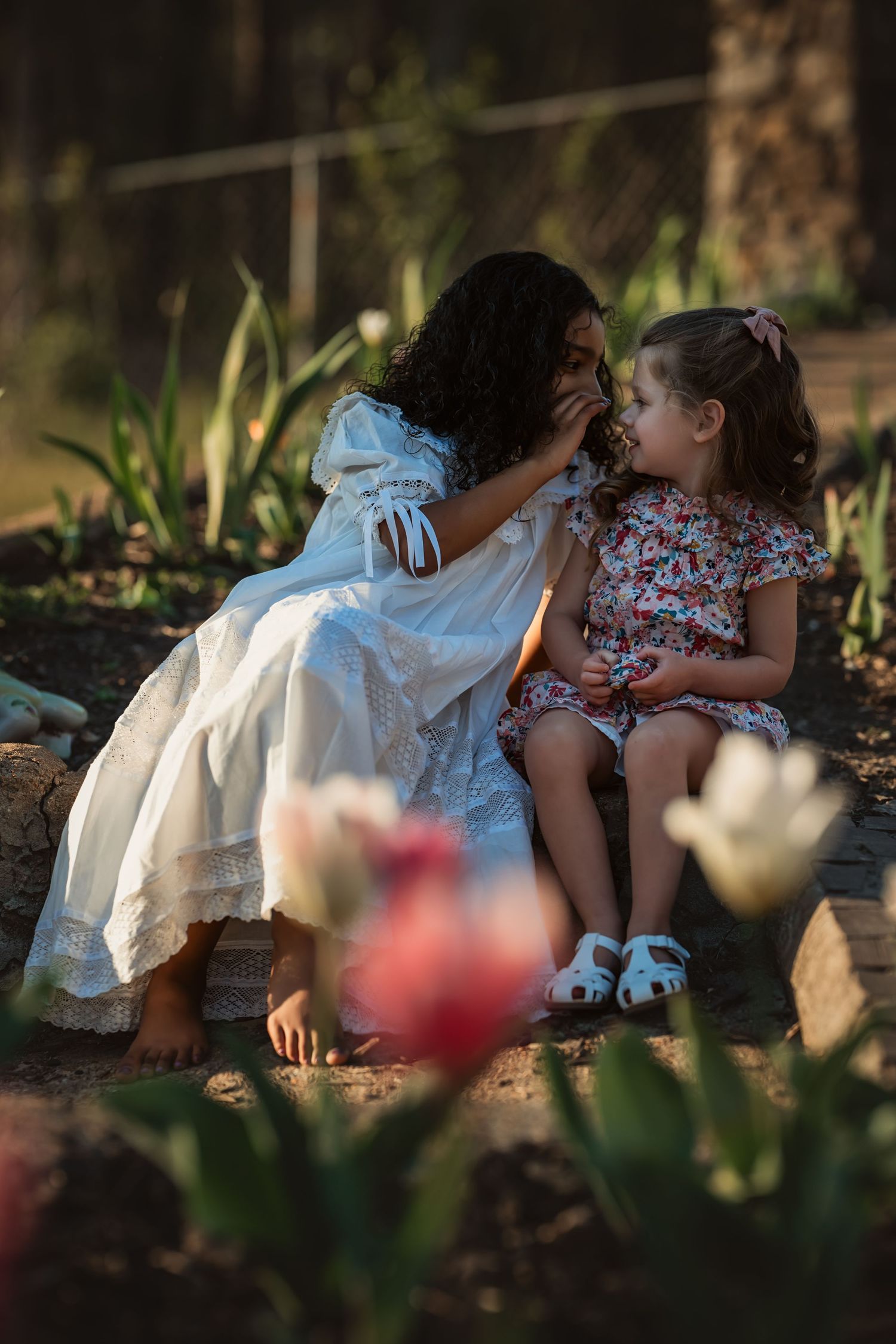 older girl in white dress whispers something to younger girl sitting on steps near tulips