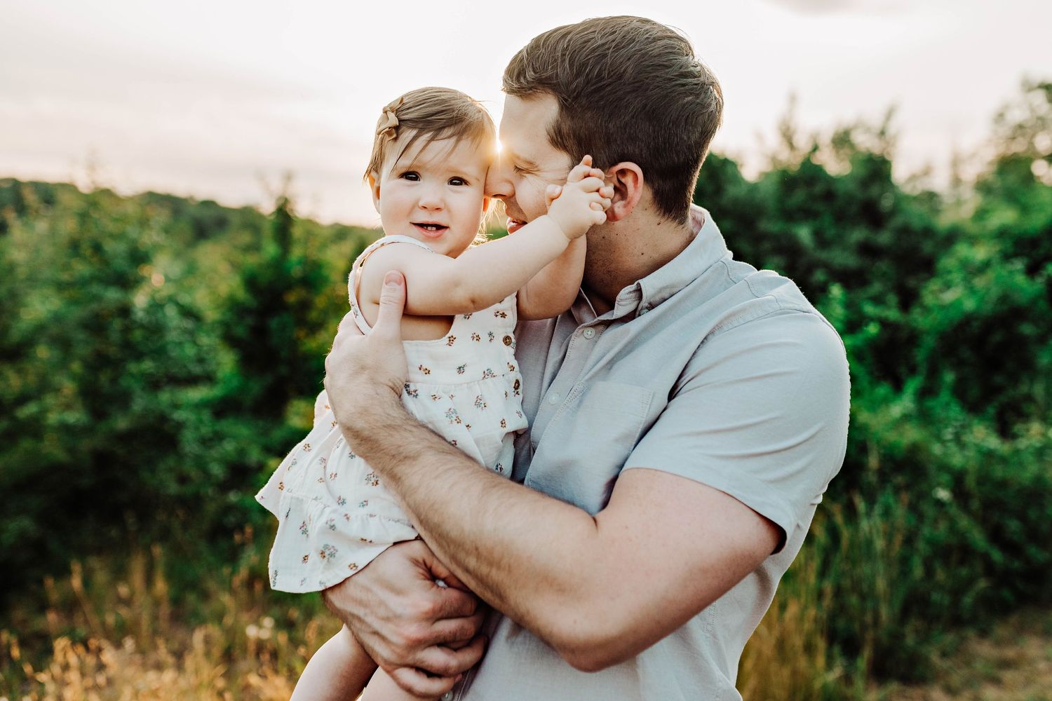 Parent and baby share an intimate outdoor moment at sunset against a green nature backdrop.