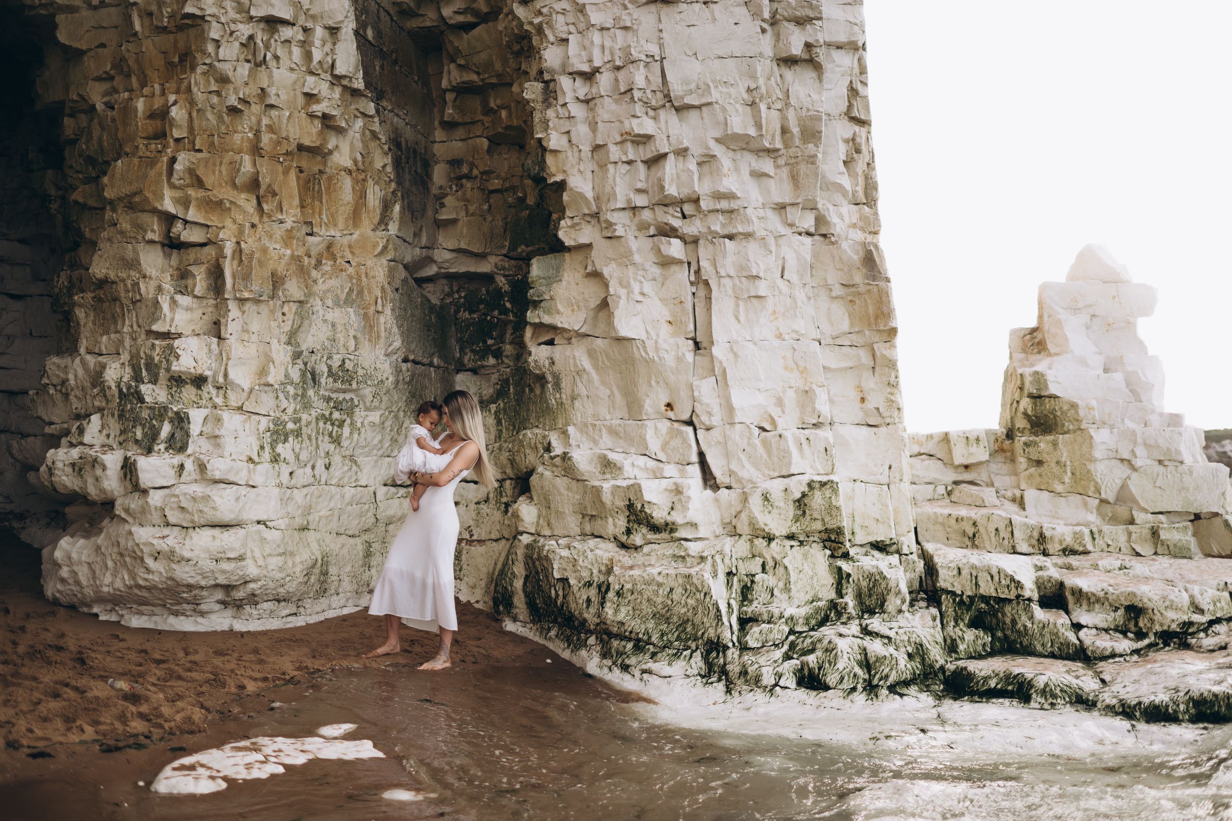 a mother with her young daughter standing near the water on a white cliff beach during a motherhood photo session in Kent