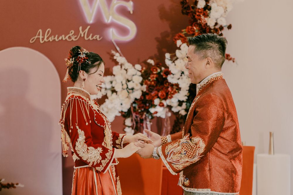 A wedding ceremony with traditional red Chinese attire against a pink backdrop with floral decorations.