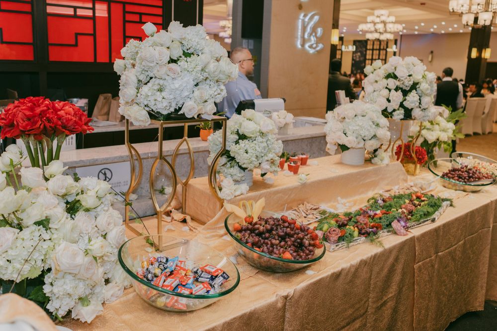 Elegant wedding reception dessert table with white and red floral arrangements, pastries, and crystal chandeliers.