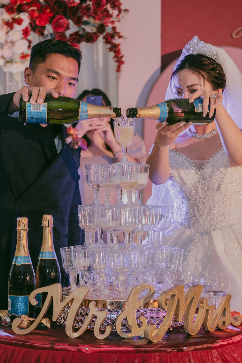Couple pouring champagne into tower of glasses at wedding reception.